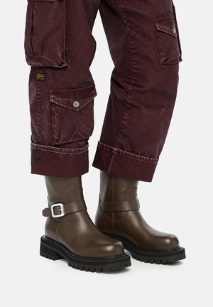 Stivaletti alla caviglia in pelle marrone con suole nere spesse e fibbie argentate, abbinati a pantaloni cargo bordeaux risvoltati.