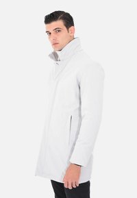 Cappotto impermeabile grigio chiaro con colletto alto, dotato di tasche laterali e una finitura liscia e testurizzata. Design slim fit con maniche lunghe.