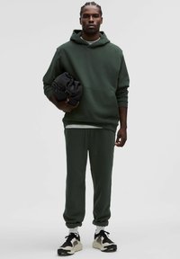 Tmavozelená mikina s kapucňou a zodpovedajúce tepláky s elastickými manžetami, doplnené svetlými teniskami. Muž drží zrolovaný čierny odev.