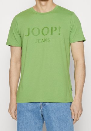 Homme portant un t-shirt vert clair à manches courtes avec le texte "JOOP! JEANS" et un jean en denim bleu, visible du cou aux hanches.