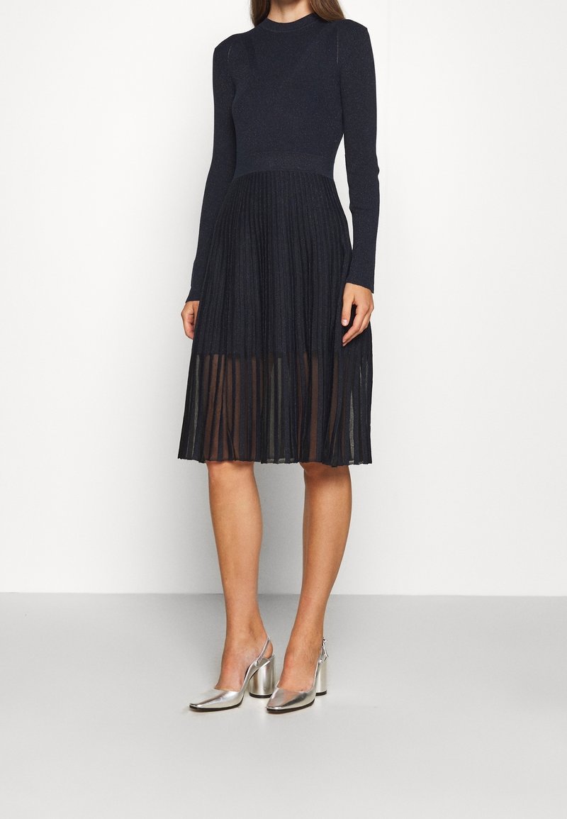 Femme portant une robe noire à manches longues avec une jupe plissée transparente et des chaussures slingback argentées à talon bloc, debout devant un fond uni.