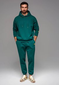 Sudadera de color verde azulado con cordones y puños acanalados, combinada con pantalones deportivos a juego, que cuentan con un parche de logo. Las zapatillas tienen un diseño en color beige y crema.