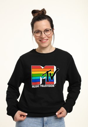 Lächelnde Frau mit Brille und Dutt, die einen schwarzen Pullover mit einem Regenbogen-gestreiften MTV Music Television Logo trägt.