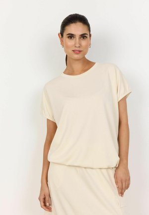 Femme aux cheveux bruns portant un haut beige ample à manches courtes et une jupe assortie, debout devant un fond blanc uni.