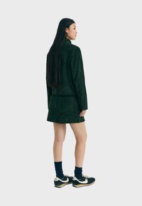 Conjunto de chaqueta de pana verde oscuro y falda mini, combinado con zapatillas negras y calcetines azules, visto desde atrás. Tela suave y texturizada.