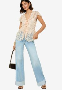 Top in pizzo rosa tenue con scollatura a V e dettagli con fiocco, abbinato a jeans a gamba larga blu chiaro con orli strappati. Borsa nera, sandali con cinturini.