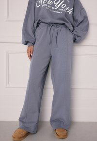 Grå sweatpants med løst fit, elastisk talje, snor og sidesømme. Båret med tan farvede slip-on sko, sat mod en neutral baggrund.