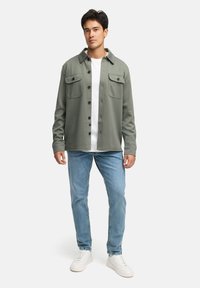 Grøn skjortejakke med knapper, to brystlommer, parret med en hvid t-shirt, lyseblå jeans og hvide sneakers, på en ensfarvet baggrund.