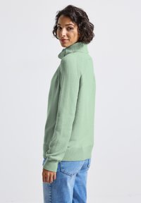 Světle zelený pletený svetr s vysokým límcem, žebrovanými manžetami a lemem. Obsahuje texturovaný vzor na ramenou a volný střih.