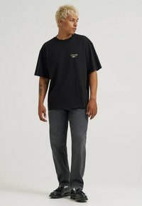 Sort oversized T-shirt med logo på venstre bryst, kombineret med grå jeans med lige ben og sorte snørebåndssko. Korte ærmer, enkelt design.