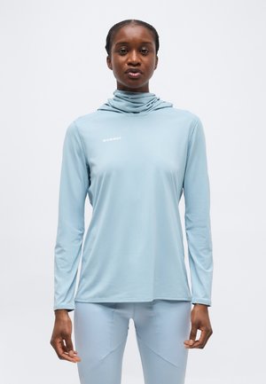 Fiatal nő világoskék hosszú ujjú felsőben és hozzáillő leggingsben, egyszerű fehér háttér előtt áll.