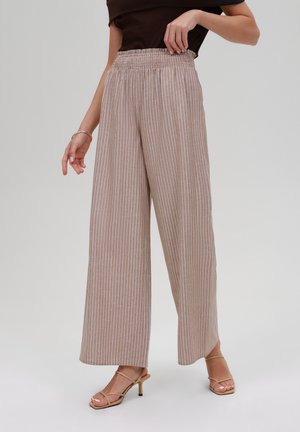 Kobieta w beżowych spodniach o szerokich nogawkach z białymi pionowymi paskami, w połączeniu z beżowymi sandałami na wysokim obcasie i czarną bluzką.