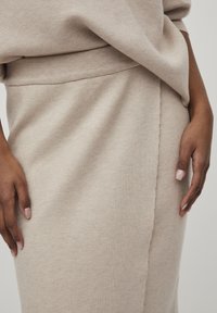 Pletená béžová sukně s upnutým pasem, hladkou texturou a viditelným švem vepředu, spárovaná s rukama jemně opřenými o látku.