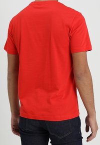 Osoba v jednoduchém červeném tričku s krátkým rukávem a tmavě modrých džínách, viděná zádama na jednobarevném světlém pozadí.