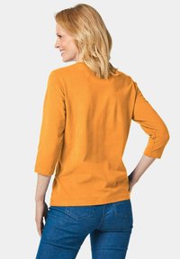 Senfgelbes T-Shirt mit 3/4-Ärmeln aus weichem Stoff, runder Ausschnitt und lockerer Passform, kombiniert mit blauen Jeans.