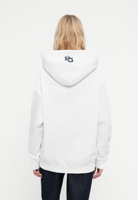 Felpa bianca con un grande cappuccio, spalle scese e un piccolo logo blu navy sul retro. Realizzata in materiale morbido con una vestibilità rilassata.