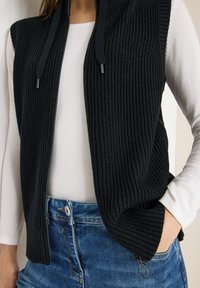 Černá pletená vesta s vertikálním žebrováním, předním zipem a kapucí. Nasažená přes bílou košili, doplněná modrými džínami.
