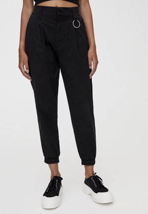 Frau trägt schwarze, hoch taillierte, zulaufende Hose mit umgeschlagenen Säumen und silbernem Ringdetail, kombiniert mit schwarz-weißen Plateau-Sneakern.