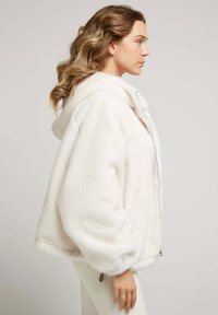 Veste à capuche en fausse fourrure blanche avec une coupe décontractée, fermeture éclair arrondie et poignets côtelés. Texture douce et manches volumineuses.