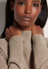 Beige Rollkragenpullover mit gerippten Bündchen, aus weich strukturiertem Stoff; getragen von einer Person, die ihren Hals hält, um Details und Wärme zu zeigen.