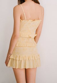 Žluté kostkované šaty vyrobené z nařasené látky, s tenkými ramínky a vrstvenými volanty na lemu. Zadní pohled ukazuje přiléhavý střih.