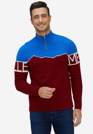 Mann lächelt, trägt einen blau-bordeauxfarbenen Zip-Up-Pullover mit weißem Text auf den Ärmeln und dunkelblaue Jeans, steht vor einem weißen Hintergrund.