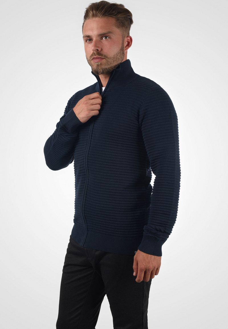 Marines zip-up sweater med tekstureret ribbestrikdesign, høj krave og lange ærmer. Bæres åben i halsen, parret med mørke bukser.
