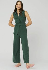 Combinaison sans manches verte avec un devant à boutons, deux poches poitrine et une ceinture nouée à la taille. Associée à des chaussures roses respirantes.