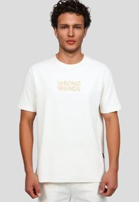 Camiseta blanca de manga corta hecha de algodón suave, con el logotipo bordado "WRONG FRIENDS" en beige en el centro. Ajuste simple y relajado.