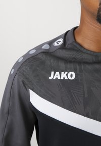 Maglietta sportiva JAKO in grigio scuro con motivo testurizzato, accenti bianchi e dettagli circolari sulle spalle. Presenta un collo rotondo e un design aderente.