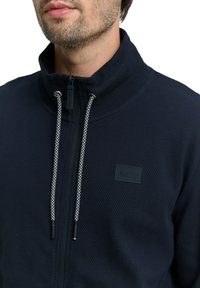 Marineblauwe jas met rits, gemaakt van een getextureerde stof, met een hoge kraag en witte gevlochten trekkoorden. Voorzien van een klein zwart logo op de borst.