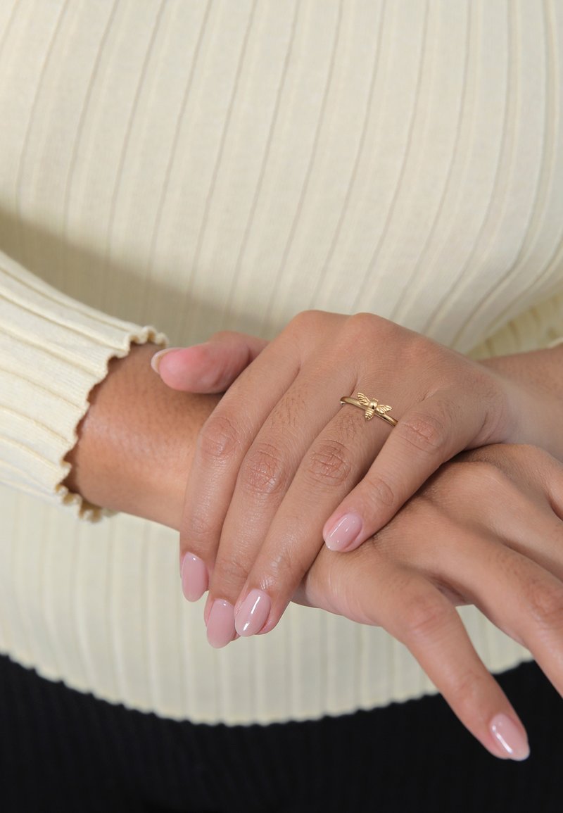 Goldring mit einem kleinen Bienenmotiv, präsentiert auf Händen mit hellrosa Nagellack, die ein cremefarbenes, geripptes Oberteil mit gewellten Ärmelabschlüssen tragen.