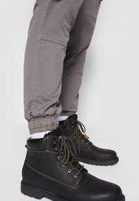 Bottines en cuir noir avec des lacets gris portées avec un pantalon de jogging gris et des chaussettes blanches sur un fond clair uni.