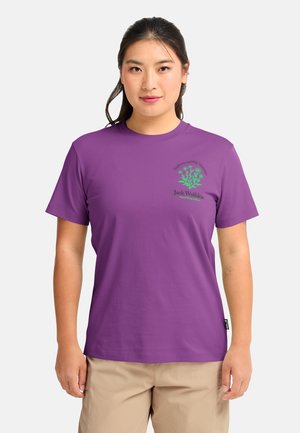 Femme portant un t-shirt violet Jack Wolfskin avec un motif floral vert et un pantalon beige, debout devant un fond clair uni.