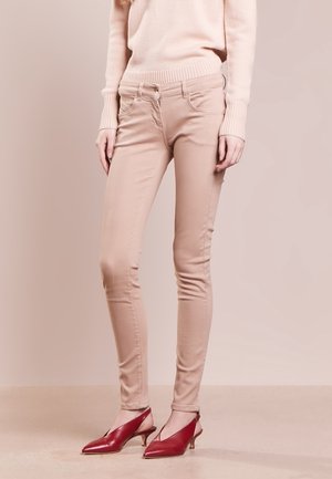 Kvinde iført beige skinny bukser, en lyserød sweater og røde spidse slingback hæle, stående mod en ensfarvet baggrund.