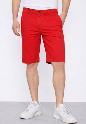 Homme portant un short rouge vif mi-long, des baskets blanches et une chemise blanche, debout avec une main dans la poche.