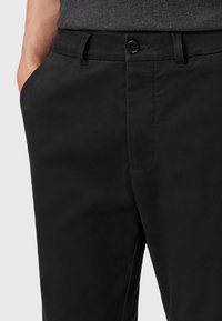 Mann mit linkem Hand in der Tasche, der schwarze knöpfbare Hosen und ein dunkelgraues Hemd vor einem schlichten Hintergrund trägt.