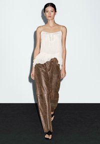 Woman walking on runway wearing sheer white sleeveless top with ruffled hem, loose brown leather pants, and black open-toe heels.