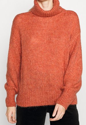 Orange turtleneck-sweater i tyk strik med lange ærmer båret af en person mod en ensfarvet baggrund.