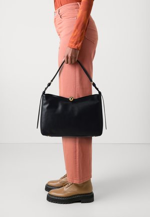 Borsa a mano in pelle nera dalla forma arrotondata e con dettagli in oro, tenuta da una persona che indossa pantaloni rosa chiaro e scarpe beige con lacci.