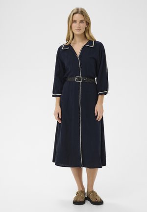 Žena stojaca v námornícky modrých midi šatách s bielym lemovaním, čiernym tkaným opaskom, trojštvrťovými rukávmi a hnedými sandálmi na bielom pozadí.