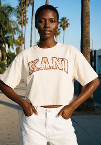 Enes kindlalt väljas, kandes lühikest beeži T-särki logoga "KANI" ja kõrge vöökohaga valgeid pükse, käed taskutes seistes.