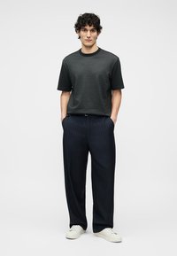 Mann med krøllete hår iført en mønstret svart kortermet skjorte, marineblå bukser med pinstripe, hvite joggesko, stående med hendene i lommene.