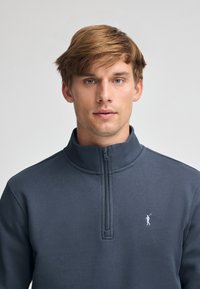 Mörkgrå fleece-pullover med halvdragkedja i kragen. Har en liten vit logotyp på vänster bröst, mjuk textur och en åtsittande design.