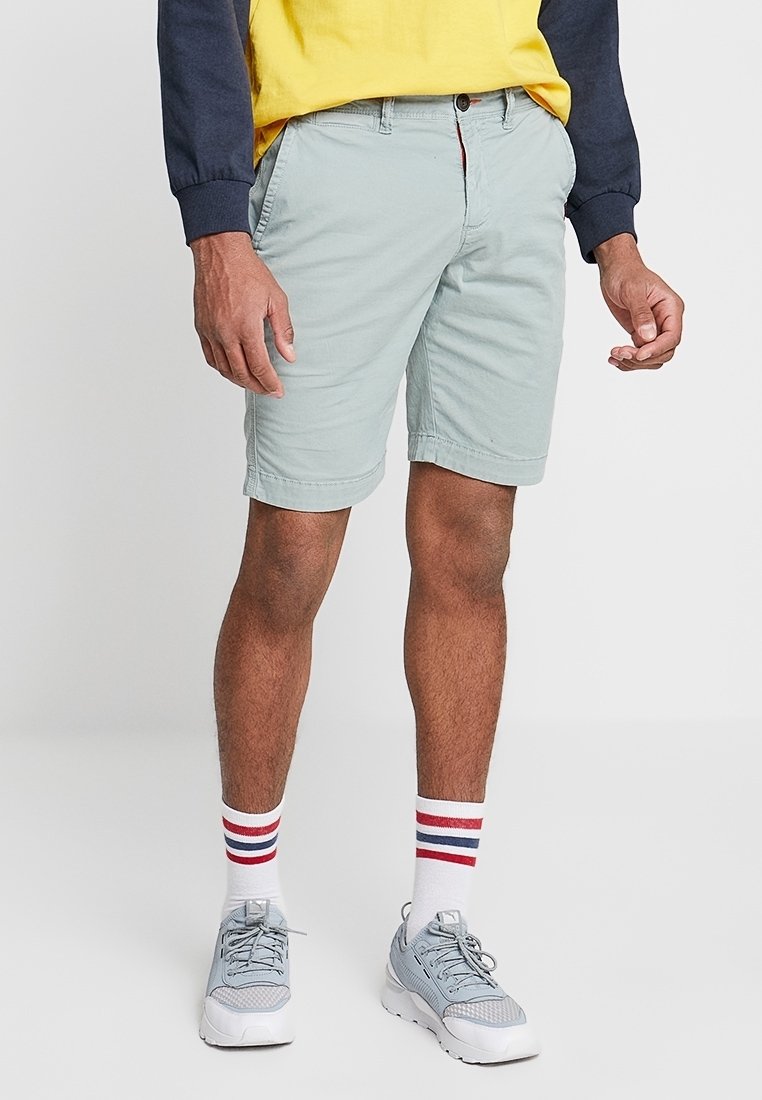 Pantalones cortos azul claro con un corte clásico, hechos de tela de algodón, con bolsillos laterales y cierre de botón. Combinados con zapatillas grises y calcetines a rayas.