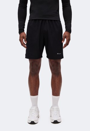 Mann trägt schwarze Sportshorts, schwarzes Langarmshirt, weiße Socken und weiße Turnschuhe, steht vor schlichtem Hintergrund.