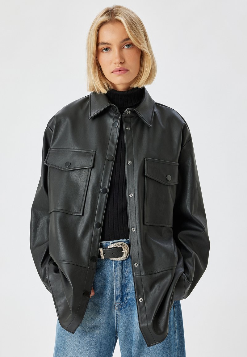 Schwarze Leder-Übergroße Jacke mit Knopfleiste, zwei Brusttaschen und glatter Textur, kombiniert mit einem schwarzen gerippten Rollkragenpullover und blauen Jeans.