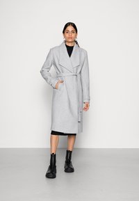 Cappotto in misto lana grigio chiaro con colletto a risvolto, cintura in vita, tasche laterali e maniche lunghe, abbinato a stivaletti neri alla caviglia.