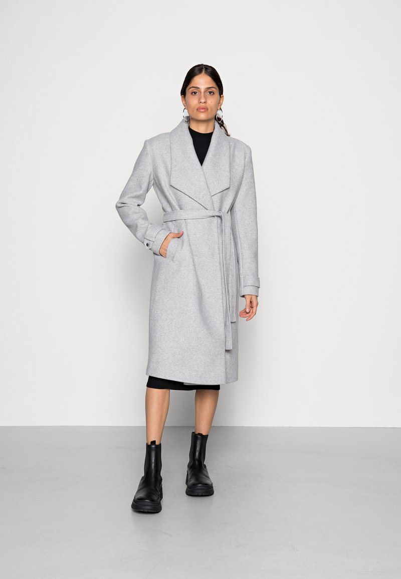 Cappotto in misto lana grigio chiaro con colletto a risvolto, cintura in vita, tasche laterali e maniche lunghe, abbinato a stivaletti neri alla caviglia.