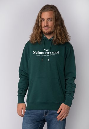 Hoodie vert foncé avec une poche avant, capuche à cordon de serrage et un graphique en texte blanc indiquant "Schau ma moi". Texture de tissu lisse avec des poignets côtelés.